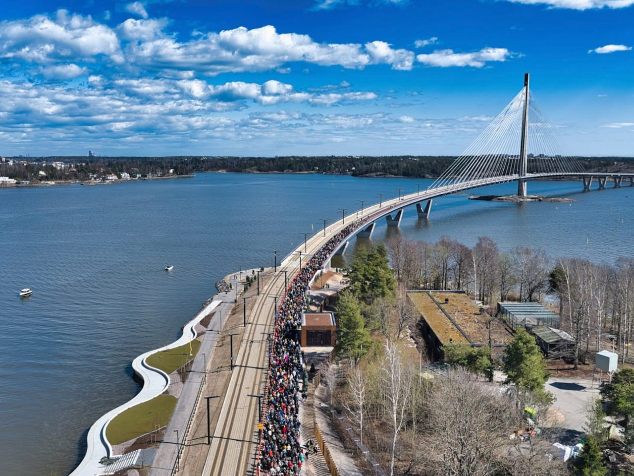 Crowd of people walking along a curved waterfront bridge over a wide river, with a cable-stayed bridge in the background under a blue sky.