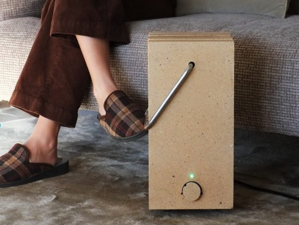 Person in plaid slippers sits on a sofa with a wooden block device on the floor, featuring a bent metal tube and a green LED light.