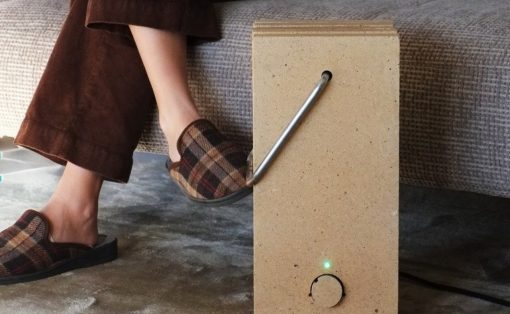 Person in plaid slippers sits on a sofa with a wooden block device on the floor, featuring a bent metal tube and a green LED light.