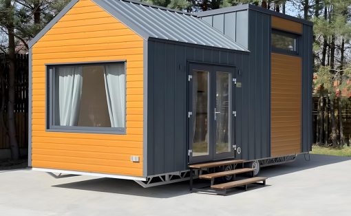 Modern tiny house on a wheeled trailer with orange and dark gray siding, large front window with white curtains, and wooden stairs to double glass doors.