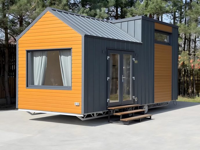 Modern tiny house on a wheeled trailer with orange and dark gray siding, large front window with white curtains, and wooden stairs to double glass doors.