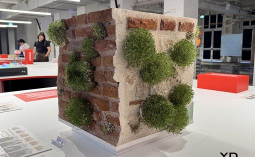 Miniature brick wall corner with beige plaster and round green topiary balls attached, on a white display table in a gallery space.