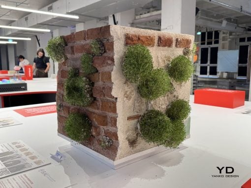 Miniature brick wall corner with beige plaster and round green topiary balls attached, on a white display table in a gallery space.