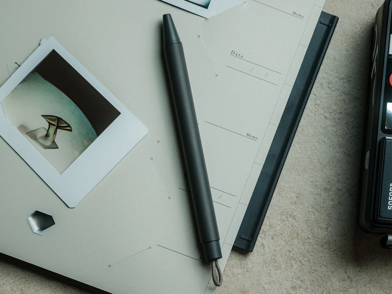 Desk scene with a black pen laid over light documents, a small Polaroid-style photo, and a calculator on a beige surface.