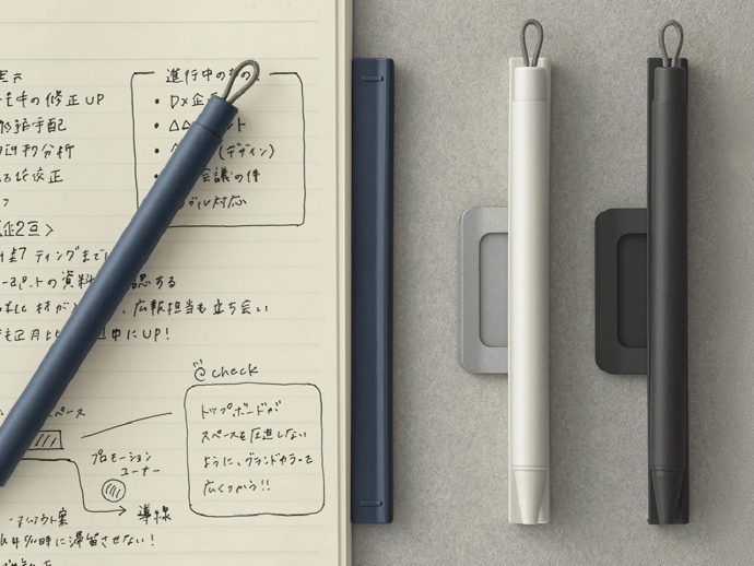 Notebook with handwritten notes in Japanese, a blue pencil resting diagonally across the page, and three pens (white, blue, black) on a gray surface.
