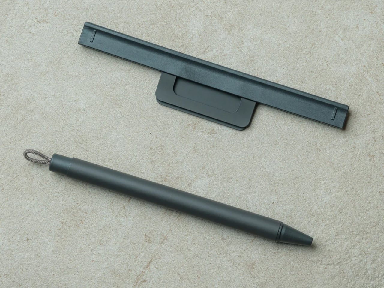Black stylus pen with a looped cord on a beige textured surface, shown beside a slim black stand or holder.