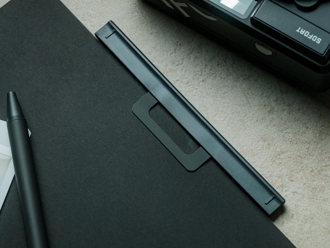 Close-up of a black notebook with a rectangular clip on its cover, a black pen nearby, and part of a camera in the corner.