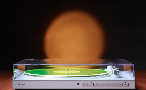 White turntable with a clear lid and a bright green vinyl on a wooden surface, orange blur in the dark background.