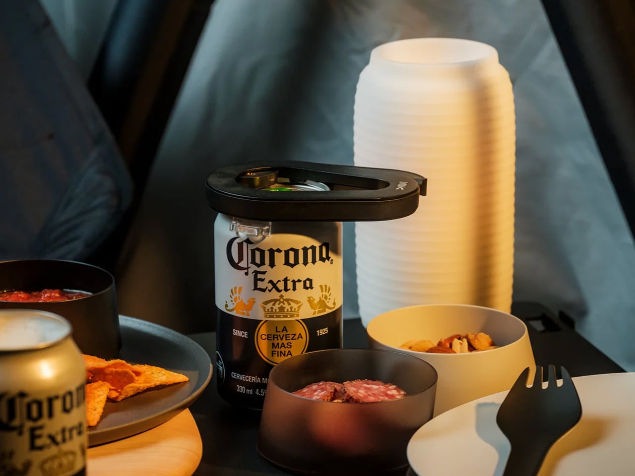 Corona Extra beer bottle at a campsite-style table with snacks, plastic cups, and a black lid or opener on top of the bottle cap