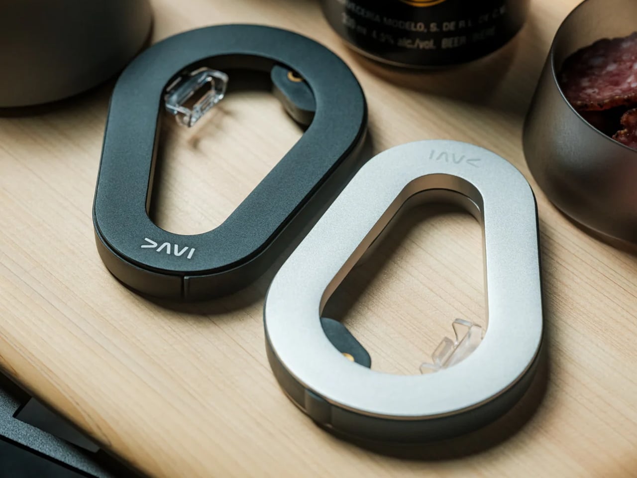 Two oval metal carabiner-style clips (one dark gray, one silver) resting on a light wood desk, with small latch mechanisms visible.