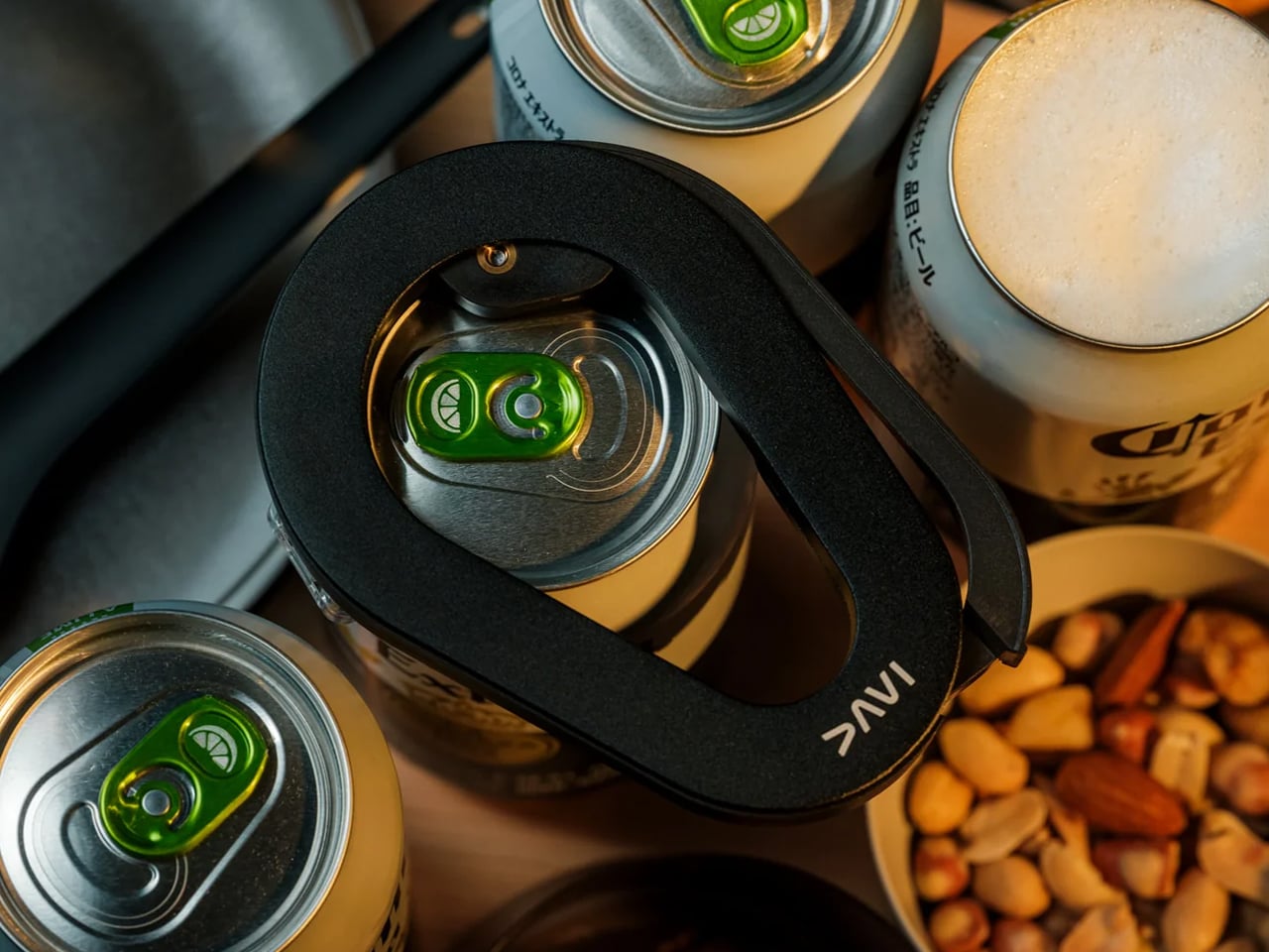Close-up of several beer cans with green pull-tabs and a black curved strap resting across the tops, a bowl of mixed nuts visible in the background.