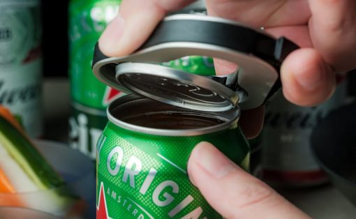 Hands lift and pull back the tab on a green beer can, revealing the opening. The can sits among other cans on a surface.