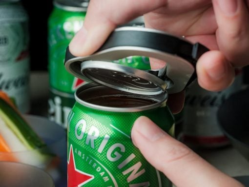 Hands lift and pull back the tab on a green beer can, revealing the opening. The can sits among other cans on a surface.