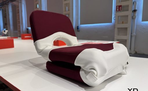 Folded maroon and white upholstered chair resting on a white display table, showroom lighting in the background, Yanko Design logo in the lower right.