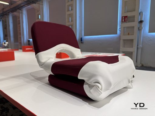 Folded maroon and white upholstered chair resting on a white display table, showroom lighting in the background, Yanko Design logo in the lower right.