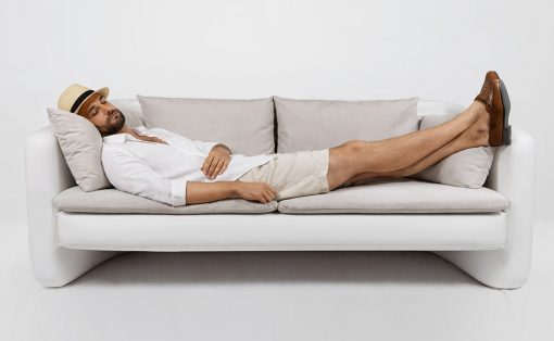 Man relaxing on a white sofa with legs propped up, wearing a hat and casual outfit.