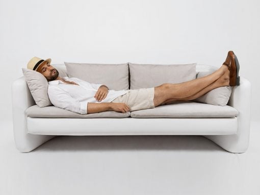 Man relaxing on a white sofa with legs propped up, wearing a hat and casual outfit.