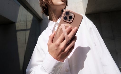 Person in a white shirt holds a pink smartphone with a circular grip against a concrete, angular architectural background.