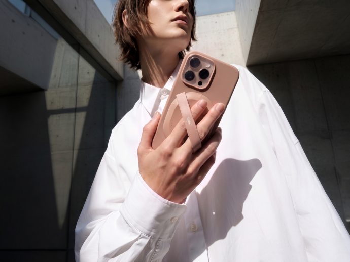 Person in a white shirt holds a pink smartphone with a circular grip against a concrete, angular architectural background.