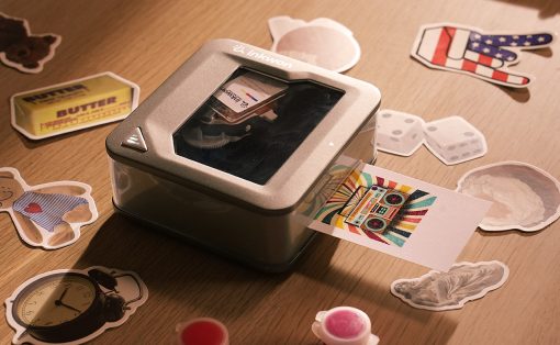 Small desktop photo printer on a wooden surface, with sticker cutouts and a colorful printed card coming out.