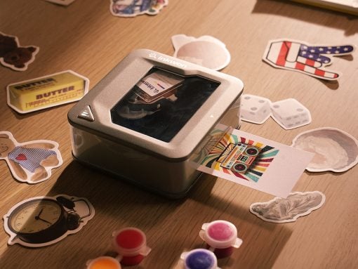 Small desktop photo printer on a wooden surface, with sticker cutouts and a colorful printed card coming out.