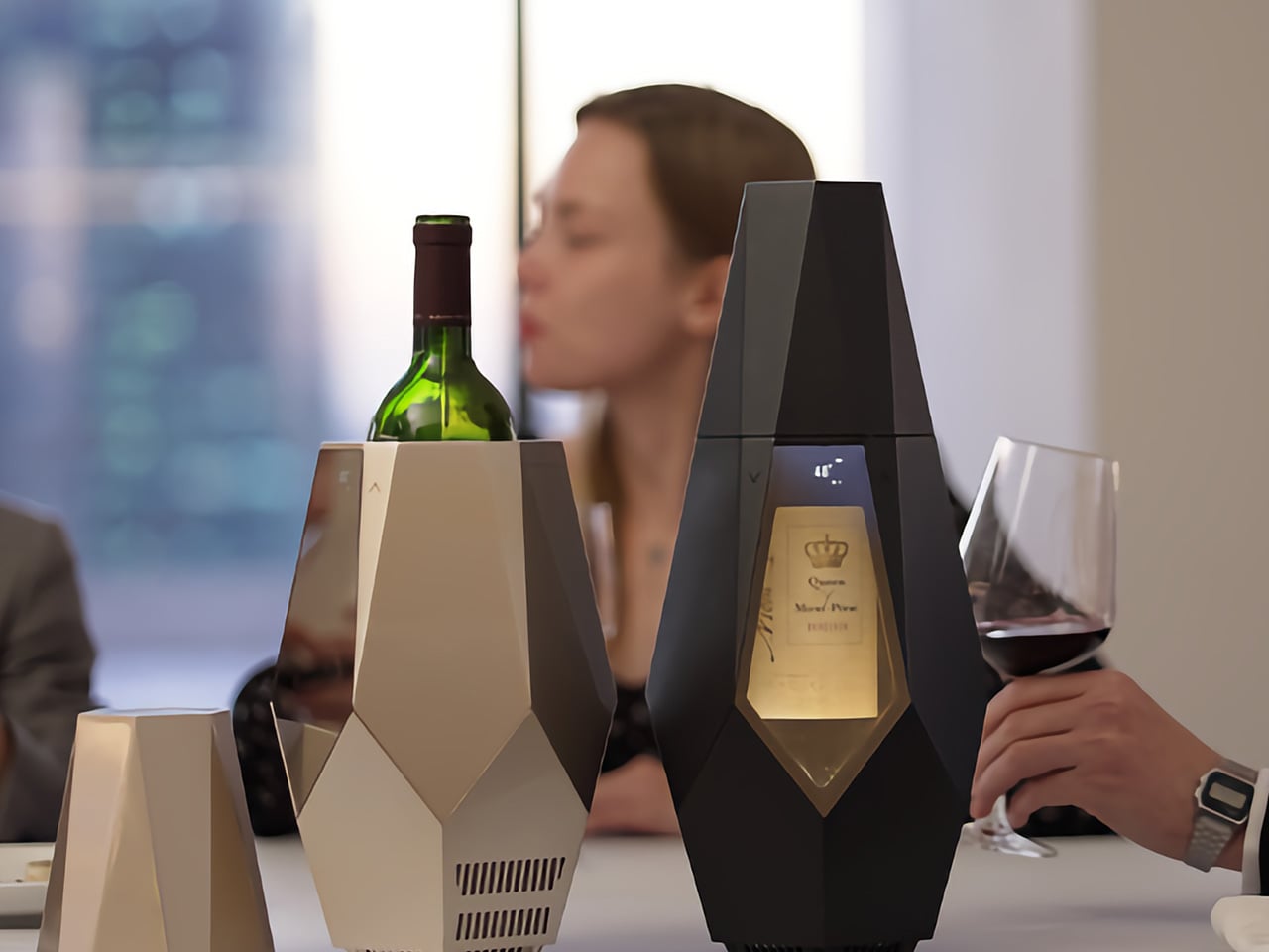 Two angular wine coolers on a table, one holding a green bottle, with a glass of red wine and a blurred person in the background.