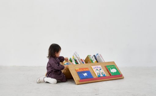 Toddler sits on the floor and browses a low wooden bookshelf filled with colorful children's books in a bright, minimal room.