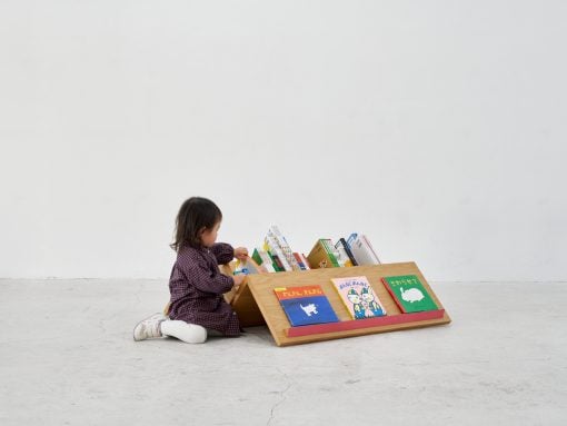 Toddler sits on the floor and browses a low wooden bookshelf filled with colorful children's books in a bright, minimal room.