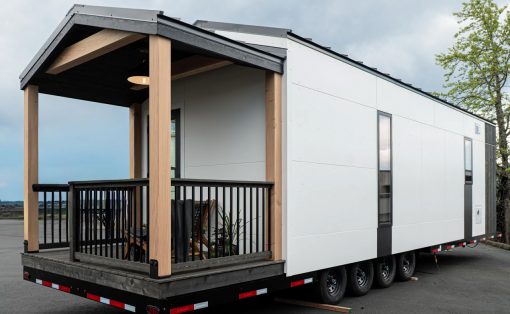 Modern tiny house on a trailer with a wooden covered porch and black railing, parked on an open lot.