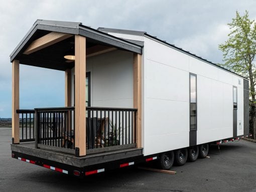 Modern tiny house on a trailer with a wooden covered porch and black railing, parked on an open lot.