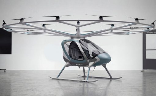Futuristic six-rotor VTOL helicopter prototype with teal accents on skids, displayed indoors in a showroom-like space