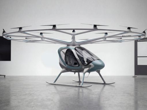 Futuristic six-rotor VTOL helicopter prototype with teal accents on skids, displayed indoors in a showroom-like space