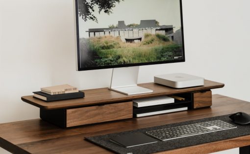 Wooden desk with an all-in-one computer monitor on a white stand, keyboard, mouse, and a minimalist desktop organizer with books and a Slim Mac Mini on the right side.