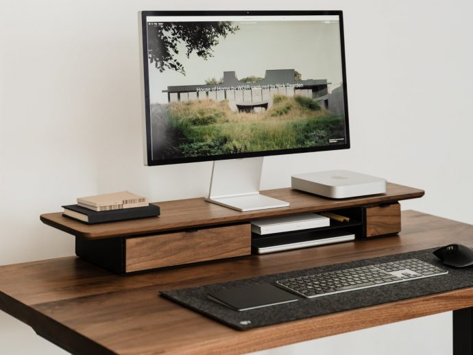 Wooden desk with an all-in-one computer monitor on a white stand, keyboard, mouse, and a minimalist desktop organizer with books and a Slim Mac Mini on the right side.