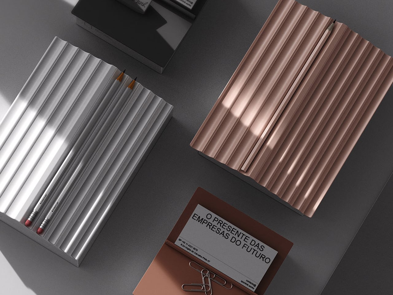 Overhead view of a dark desk with two ribbed metal organizers in silver and rose gold, plus a brown brochure with paper clips and pencils nearby in a modern setup.