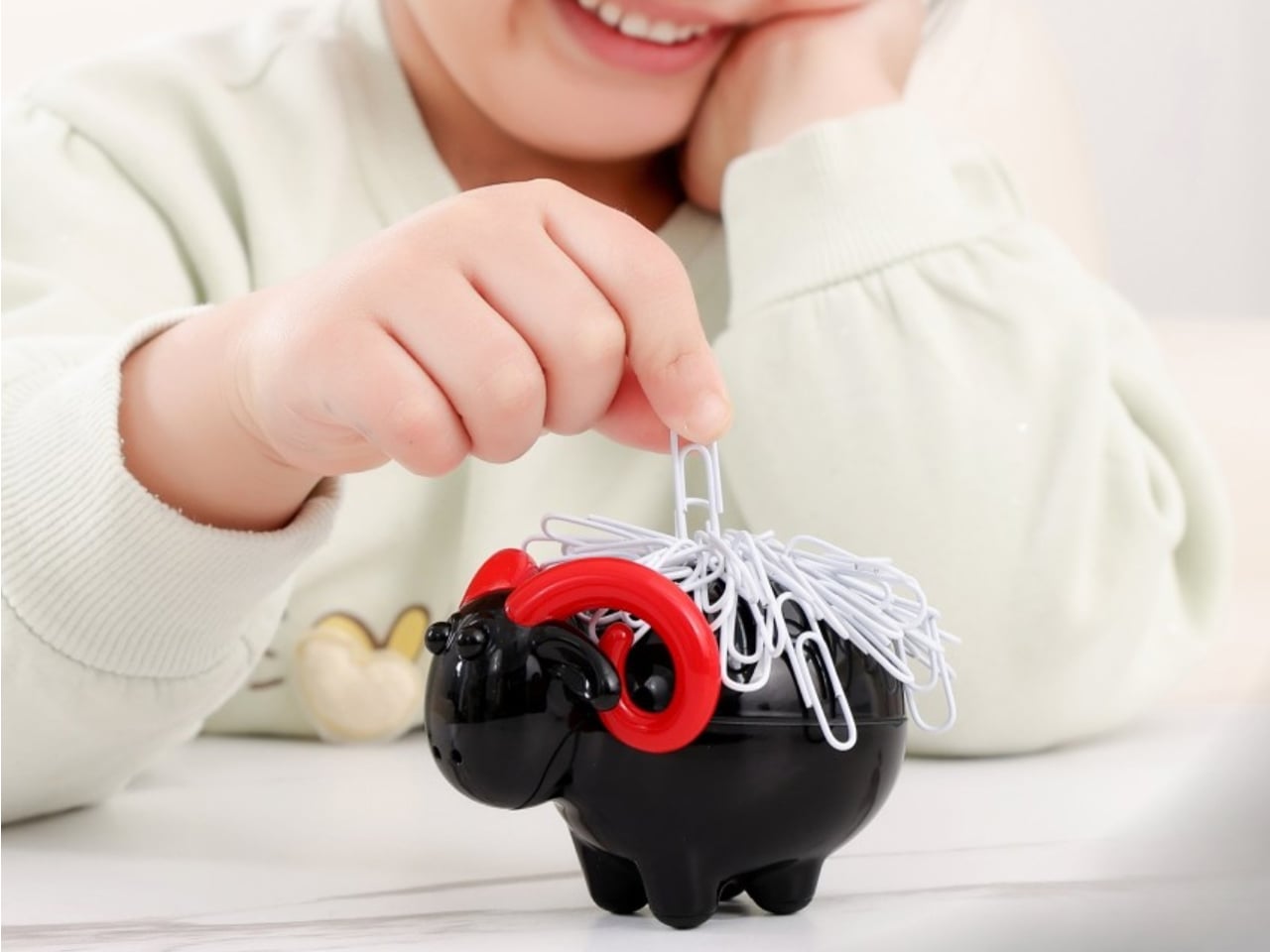Child smiling while threading a paperclip into a black piggy bank with a tangled nest of white clips on top