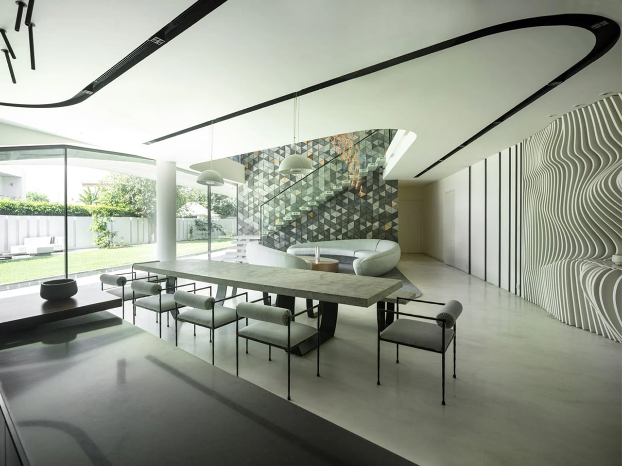 Open-plan dining area with a long marble table and modern chairs, glass walls overlook a green yard, geometric wall behind a curved sofa.