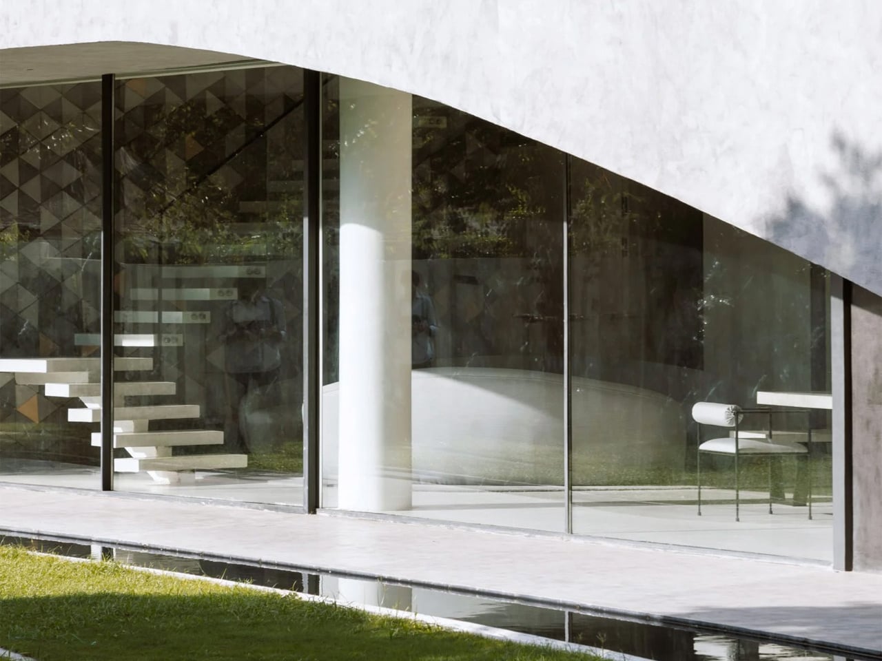 Modern glass-walled room with a white floating staircase and a minimalist chair, reflecting trees outside.
