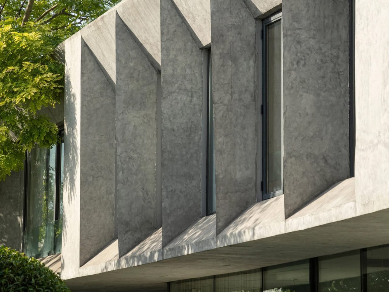Modern concrete building with angled vertical bays and tall narrow windows, shaded by a tree on the left.