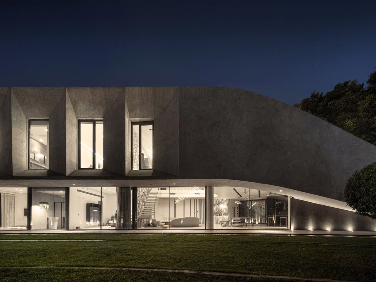 Night view of a modern concrete house with a curved roof and large glass walls revealing a warmly lit living space inside.