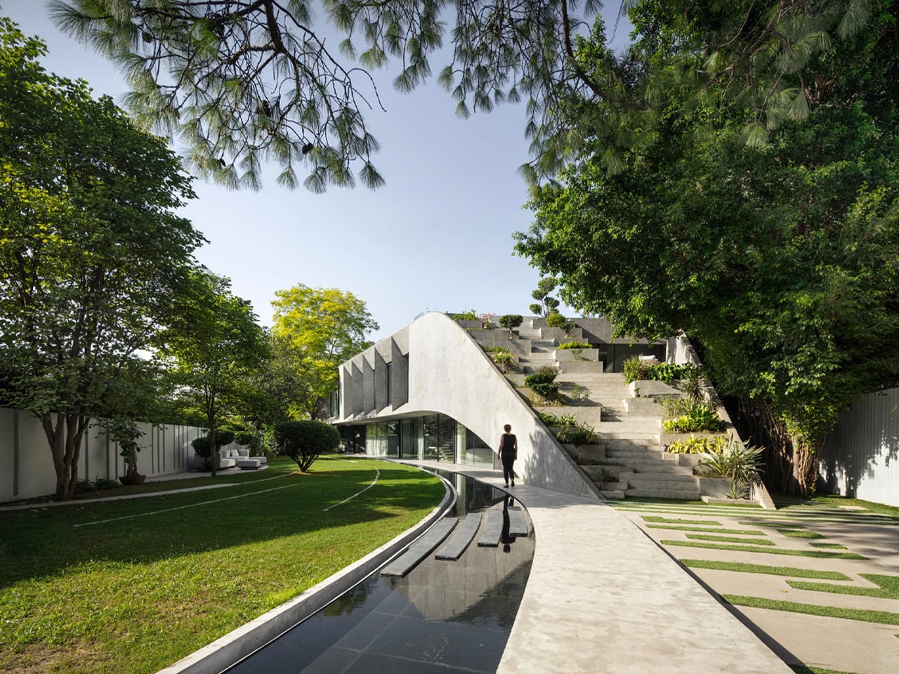 Curved concrete walkway beside a narrow reflective canal leads to a modern terraced garden with trees and lawn.