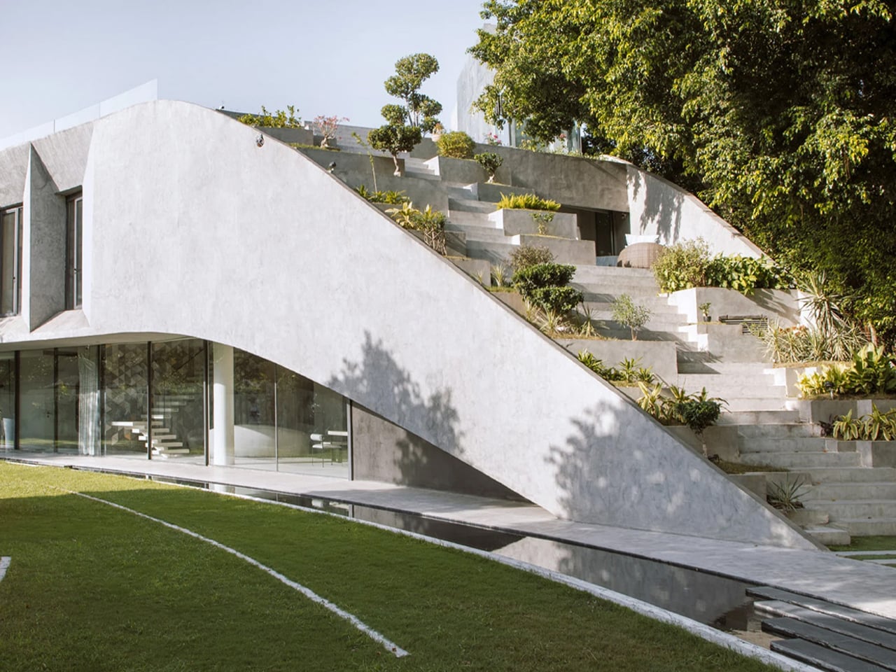 Modern white concrete house with a curved ramp and terraced garden surrounded by trees.