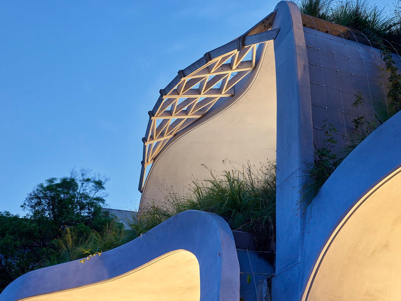 Curved blue concrete wall with a lattice window, illuminated by warm lights at twilight.