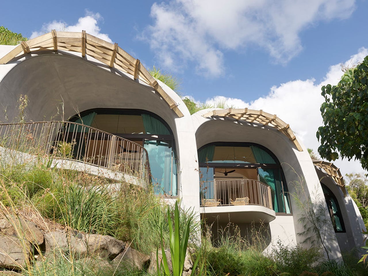 Curved modern hillside house with large glass doors, balconies, and a wooden pergola on top, surrounded by tall grasses and rocks.