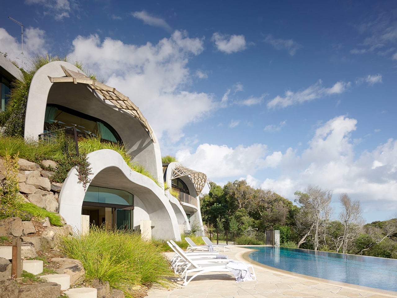 Architectural hillside villa with curved concrete arches, glass doors, and a poolside deck with white lounge chairs under a blue sky.