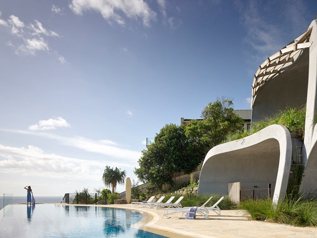 Person standing at the edge of an infinity pool, gazing toward the sea, with modern curved concrete architecture in the background.