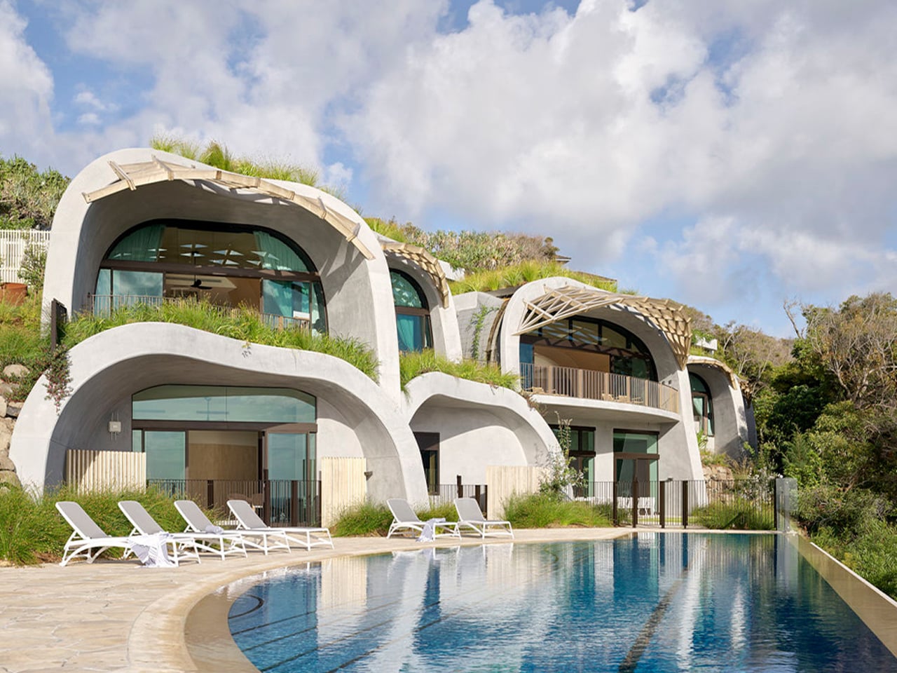 Contemporary curved concrete villas with greenery on a hillside and a large outdoor pool with white loungers in the foreground at a resort.