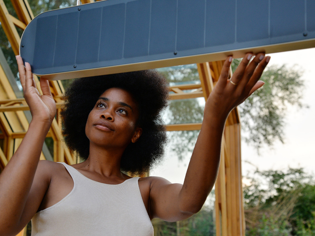 Woman outdoors lifting a blue panel of a playground structure above her head, wearing a white tank top and looking up thoughtfully.