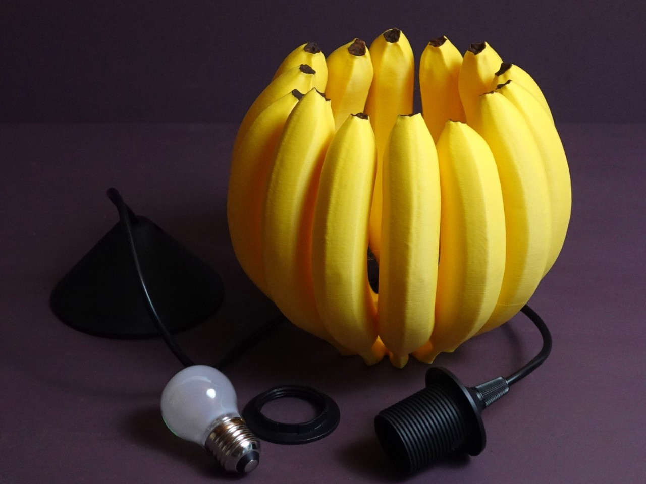 Yellow banana-shaped lamp sculpture formed by curved bananas, with a bulb and socket visible on a dark background.