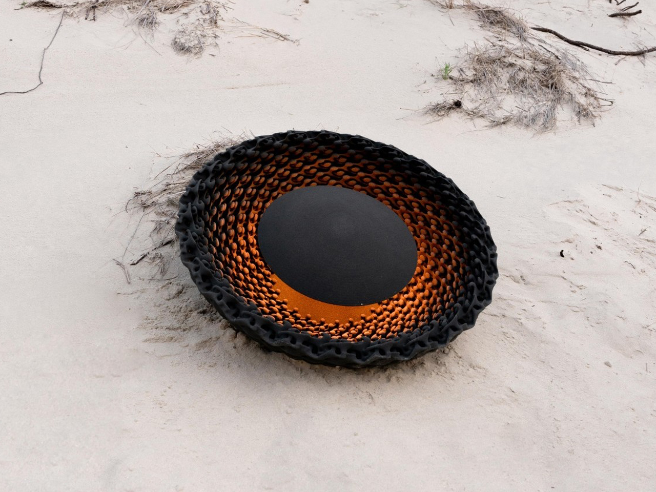 Circular black-and-orange woven sculpture resting on light beach sand.